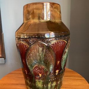 Può includere: Un vaso decorativo in ceramica con una base larga e un collo più stretto. Il vaso presenta una combinazione di colori marrone, verde e rosso con un design strutturato e fantasia. Il vaso è appoggiato su un tavolo di legno.