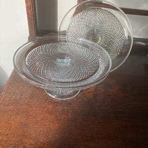 May include: A clear glass cake stand with a pedestal base and a matching lid. The stand and lid feature a textured, swirling pattern. The stand is displayed on a dark wooden surface.