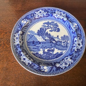 May include: A blue and white ceramic plate with a rural landscape design. The plate's central image depicts a village scene with houses and trees, framed by a floral border. The plate is a decorative item.