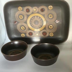 Op de afbeelding: Een bruine keramische serveerschaal en twee bijpassende kommen. De schaal heeft een rechthoekige vorm met een decoratief patroon van concentrische cirkels in bruin, beige en goudtinten. De kommen zijn rond en hebben een vergelijkbare bruine kleur.