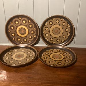 May include: Four brown ceramic plates featuring a circular floral design in gold, tan, and olive green. The plates have a dark brown border and are displayed on a wooden surface.