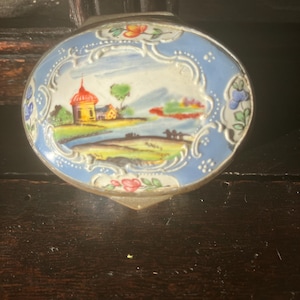 Può includere: Piccola scatola decorativa ovale con una scena di paesaggio dipinta. La scatola ha un bordo azzurro con decorazioni floreali e una cornice in metallo color argento. Il paesaggio raffigura un edificio giallo con tetto rosso, alberi e acqua.