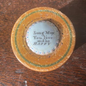 May include: A circular wooden trinket box with a painted gold and green border. The centre has a white ceramic insert with the words "Long May You live and be HAPPY" in black lettering, surrounded by blue and pink dots.