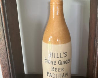 May include: A vintage stoneware bottle with a tan top and off-white base. The bottle is labeled "Hill's Stone Ginger Beer Padiham" in black lettering. The bottle is displayed on a dark wooden shelf.