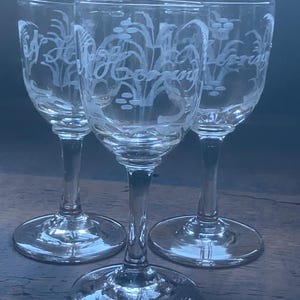 Op de afbeelding: Drie heldere likeurglazen met geëtste bloemen- en scriptontwerpen. De glazen hebben een slanke steel en een ronde voet. Het geëtste ontwerp lijkt "Morning" te lezen. De glazen zijn gerangschikt op een donker houten oppervlak.