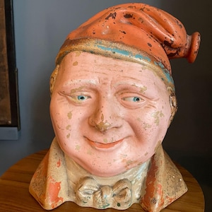 May include: A vintage, hand-painted bust of a smiling man with blue eyes, wearing an orange cap with a blue stripe and a bow tie. The bust has a pink face and a cream-coloured collar, with visible signs of wear and tear.