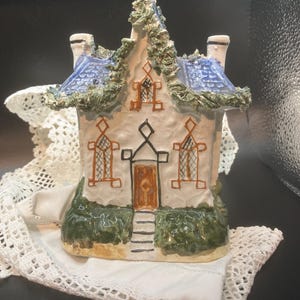 Könnte beinhalten: Eine Keramikhausfigur mit blauem Dach, grünen Verzierungen und einer braunen Tür. Das Haus hat zwei Schornsteine und dekorative Fenster. Die Figur steht auf einem weißen, mit Spitze besetzten Tuch.