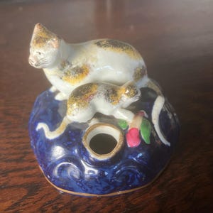 Op de afbeelding: Een keramieken kattenbeeldje met een wit en bruin gevlekte kat bovenop een donkerblauwe basis. De basis heeft een gat en is versierd met krullen en een gouden rand. Er staat ook een kleinere kat op de basis.