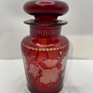 Könnte beinhalten: Eine rubinrote Glasflasche mit passendem Verschluss. Die Flasche hat geätzte Trauben- und Blattmotive. Der Verschluss hat eine abgerundete Form. Die Flasche hat ein dekoratives wellenförmiges Design um den Hals.