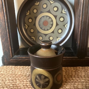 Może przedstawiać: Brązowy ceramiczny słoik z pokrywką, z kwiatowym wzorem w kolorze żółtym i brązowym. Pasujący talerz z podobnym wzorem znajduje się w tle. Słoik stoi na tkanej macie, tworząc estetykę vintage.