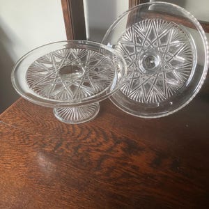 May include: Two clear glass cake stands with starburst patterns. One stand is elevated on a pedestal base, while the other is a flat plate. Both have a decorative, textured design, perfect for displaying desserts or other items.