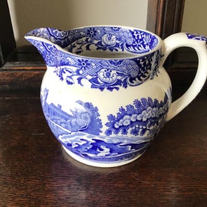 Könnte beinhalten: Eine weiß-blaue Keramikkrug mit Landschaftsmuster. Der Krug hat einen abgerundeten Körper, einen gebogenen Griff und einen Ausguss. Das blaue Design umfasst florale und szenische Elemente. Der Krug ist wahrscheinlich Vintage oder antik.