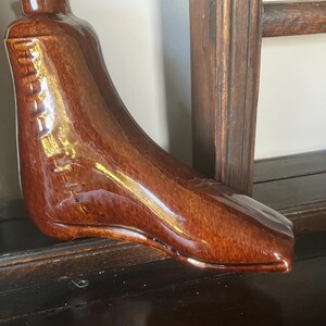 Op de afbeelding: Een bruine keramische fles in de vorm van een laars met een glanzende afwerking. De fles heeft een gedetailleerd ontwerp, inclusief stikseldetails aan de zijkant. Het is een decoratief item.