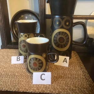 May include: A set of vintage brown ceramic mugs and a pitcher with a floral design in yellow and orange. The pitcher and mugs have a dark brown handle and a white interior. The items are arranged on a woven mat.