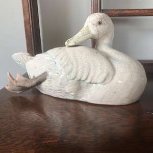 May include: A white ceramic duck figurine with a visible crackle glaze finish. The duck is depicted in a resting position with its head turned toward its back and one webbed foot extended outward. The surface features subtle blue-toned highlights.