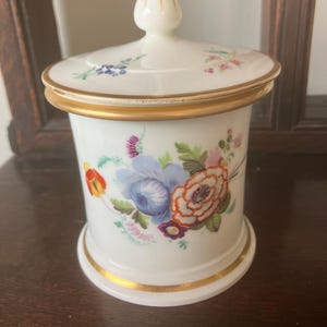 May include: A white porcelain jar with a lid, adorned with colorful floral designs. The jar features a gold rim and base, and the lid is topped with a gold-tipped finial. The floral pattern includes blue, orange, and pink flowers.