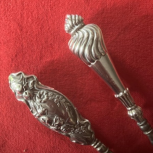 Könnte beinhalten: Zwei silberfarbene Metallbestecke mit verzierten Griffen. Ein Griff zeigt ein detailliertes geprägtes Blumenmuster mit Blättern und Blüten, während der andere Griff ein glattes, spiralförmiges und geripptes Design mit einer spitzen Endkappe aufweist.