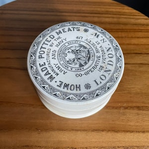May include: A vintage, round, white container with a black and white patterned design. The lid features text that reads "Potted Meats" and "Home Made." The container is from the Army & Navy Co-operative Society, London.