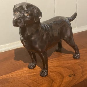 Puede incluir: Figura de cerámica negra brillante de un Labrador Retriever. El perro está de pie, con la cola ligeramente levantada. La figura tiene detalles, incluyendo ojos, nariz y patas. El perro está sobre una superficie de madera.