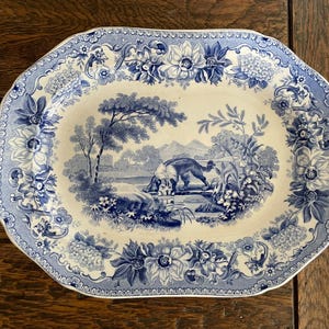 May include: An oval ceramic platter featuring a blue and white transferware pattern. The center depicts a dog near a body of water with trees and hills. The wide border is decorated with detailed floral motifs including large blossoms.