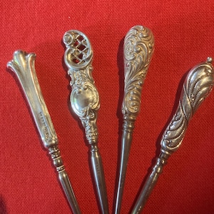 Könnte beinhalten: Vier silberfarbene Metallwerkzeuge mit verzierten, dekorativen Griffen. Jeder Griff weist ein einzigartiges geprägtes Muster auf, darunter florale, gitterartige und geschwungene Designs. Die Werkzeuge haben lange, schlanke, glatte Schäfte.