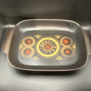 May include: A rectangular, dark brown baking dish with a decorative floral pattern in gold, orange, and brown. The dish has two handles and is suitable for oven use. The design is centered on the dish.