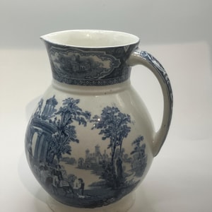 Może przedstawiać: Biały ceramiczny dzban z niebiesko-białą sceną pasterską. Dzban ma zaokrąglony korpus, dzióbek i zakrzywiony uchwyt. Obręcz i uchwyt są ozdobione niebieskimi wzorami.