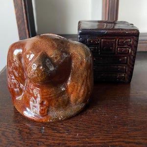 May include: A glossy brown ceramic figurine shaped like a curled animal sits beside a dark brown, square ceramic box featuring a molded grid pattern on its front face that resembles a small chest of drawers with multiple compartments.
