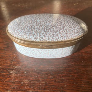 May include: An oval-shaped trinket box with a light blue speckled lid and sides, accented with a gold-colored metal rim. The box sits on a dark wooden surface, showcasing its vintage aesthetic.