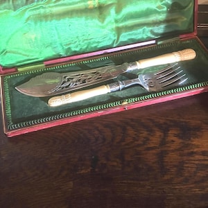 Puede incluir: Un juego de servir pescado plateado que consta de un cuchillo grande y un tenedor con patrones grabados ornamentados en las hojas y dientes de metal. Los mangos son de color blanco roto con detalles tallados, presentados en un estuche rojo.