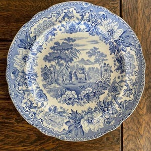 May include: A round ceramic plate featuring a blue and white transferware pattern. The center depicts a pastoral landscape with sheep and trees, while the wide border is decorated with detailed floral motifs and scrolling foliage.