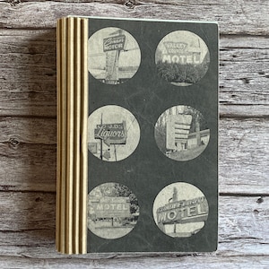 May include: A vintage-style journal with a dark gray cover featuring six circular black and white images of retro motel and liquor store signs. The spine is a textured gold color. The journal is likely a travel or memory-keeping item.