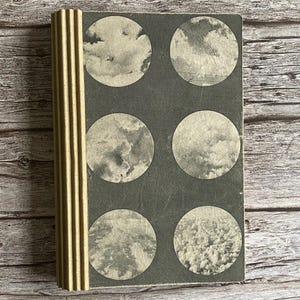 May include: A vintage journal with a dark gray cover featuring six circular cloudscape images. The spine is a corrugated gold color. The journal is open on a wooden surface.