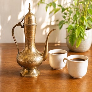 May include: A vintage brass coffee pot with intricate floral patterns, a long spout, and a curved handle. Two white ceramic cups filled with dark coffee sit beside the pot. A potted plant is in the background.