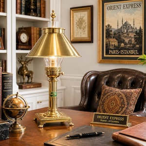 Op de afbeelding: Een messing bureaulamp met een conische kap en een heldere glazen lamp. De lamp staat op een houten bureau met een globe, boeken en een bord met de tekst "ORIENT EXPRESS PARIS-ISTANBUL". Een ingelijste poster is zichtbaar op de achtergrond.