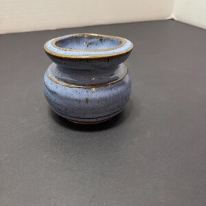 May include: A small, handcrafted ceramic vase in a light blue hue with brown trim. The vase has a rounded body and a flared rim, showcasing a simple yet elegant design. The pottery appears to be glazed, giving it a smooth finish.