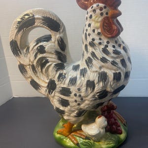 Pode incluir: Estatueta de galo de cer&acirc;mica com corpo salpicado de branco e preto, crista e barbela vermelhas e patas laranja. O galo est&aacute; sobre uma base verde com um pintinho branco e uvas roxas. Um item decorativo para cozinha ou casa.