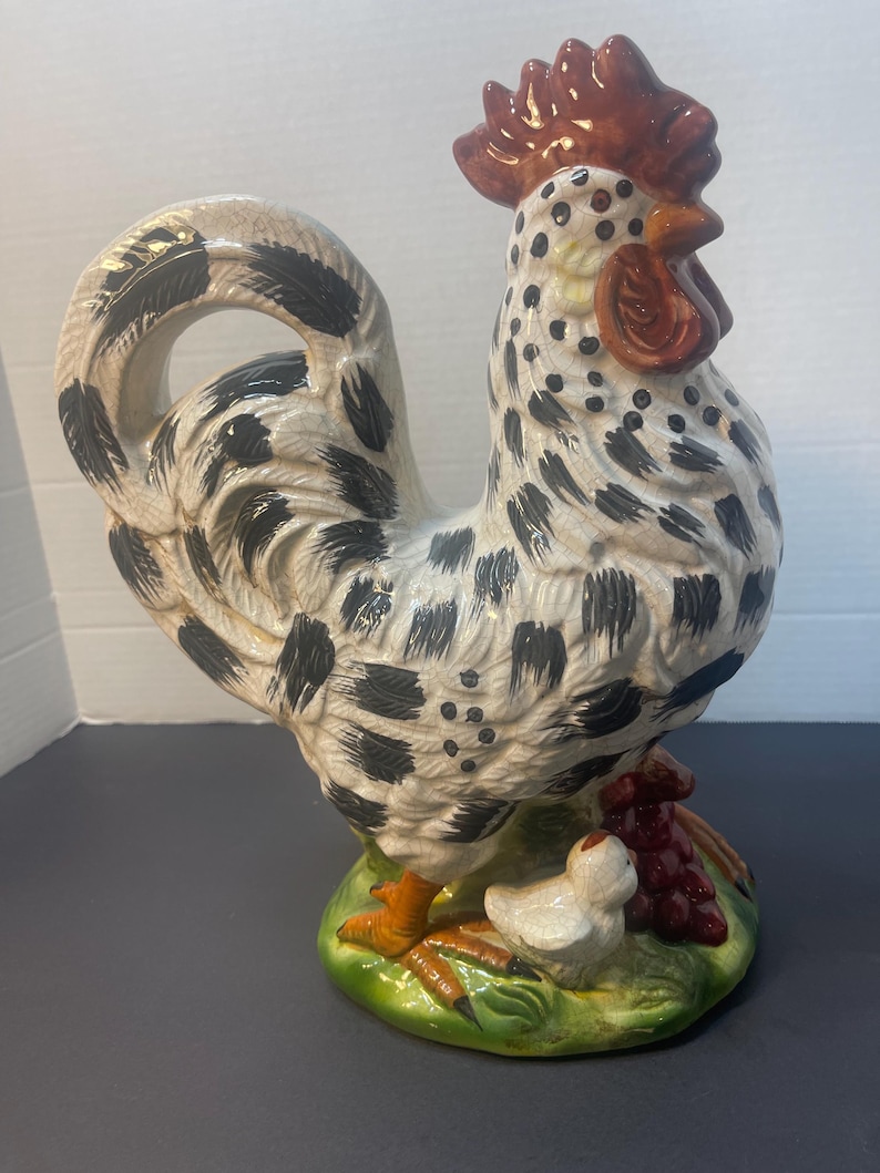 Pode incluir: Estatueta de galo de cer&acirc;mica com corpo salpicado de branco e preto, crista e barbela vermelhas e patas laranja. O galo est&aacute; sobre uma base verde com um pintinho branco e uvas roxas. Um item decorativo para cozinha ou casa.