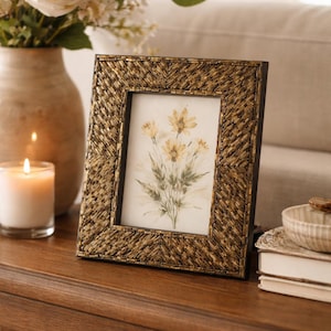 May include: A decorative framed picture with a gold beaded border, displaying a watercolor floral print. The frame sits on a wooden surface, next to a lit candle and a vase of flowers. The overall theme is home decor.