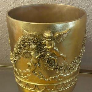May include: A gold-colored decorative vase with cherub and floral relief details. The vase features two cherubs with wings, surrounded by roses and clouds. The vase has a rounded base and a wide opening.