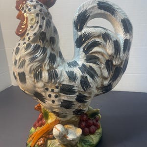 Pode incluir: Estatueta de galo de cer&acirc;mica com corpo salpicado branco, preto e cinza, crista e barbela vermelhas e cauda curva. A base apresenta uvas e um p&aacute;ssaro pequeno. O galo &eacute; um item decorativo.
