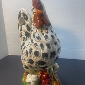 Pode incluir: Estatueta de galo de cer&acirc;mica com corpo branco salpicado de manchas pretas. O galo tem crista e barbela vermelhas e est&aacute; sobre uma base verde com um cacho de uvas vermelhas e dois pintinhos.