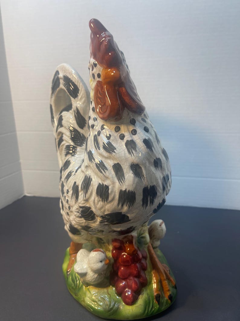 Pode incluir: Estatueta de galo de cer&acirc;mica com corpo branco salpicado de manchas pretas. O galo tem crista e barbela vermelhas e est&aacute; sobre uma base verde com um cacho de uvas vermelhas e dois pintinhos.