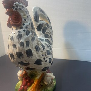 Pode incluir: Estatueta de galo de cer&acirc;mica com corpo salpicado de branco e preto, crista e barbela vermelhas e patas laranja. O galo est&aacute; sobre uma base verde com uvas vermelhas e pintinhos. Um item decorativo.