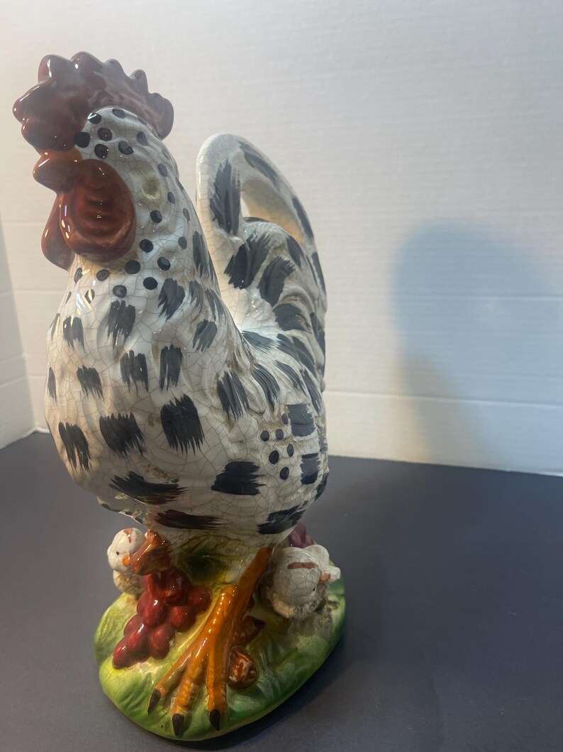 Pode incluir: Estatueta de galo de cer&acirc;mica com corpo salpicado de branco e preto, crista e barbela vermelhas e patas laranja. O galo est&aacute; sobre uma base verde com uvas vermelhas e pintinhos. Um item decorativo.