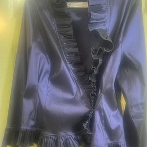 May include: A dark navy blue jacket with a ruffled collar and hem. The jacket has long sleeves and a shiny, satin-like finish. The ruffles are a darker shade of navy blue.