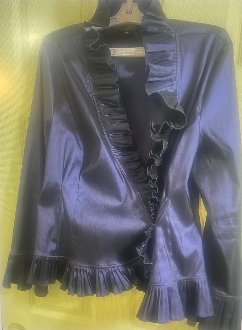 May include: A dark navy blue jacket with a ruffled collar and hem. The jacket has long sleeves and a shiny, satin-like finish. The ruffles are a darker shade of navy blue.