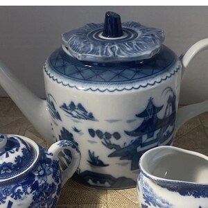 Può includere: Servizio da tè in porcellana con un design paesaggistico blu e bianco. Il set include una teiera con coperchio floreale, una zuccheriera e una lattiera. La teiera ha un lungo beccuccio e un manico curvo. Zuccheriera e lattiera presentano motivi blu e bianchi coordinati.