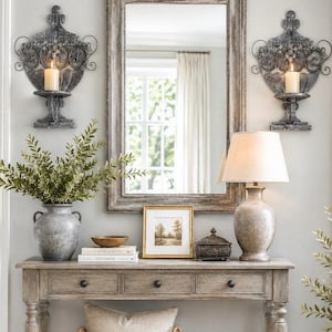 Puede incluir: Una mesa consola de madera con tres cajones, coronada por un espejo rectangular, dos apliques de pared de metal ornamentado con velas, un jarrón de cerámica con follaje, una lámpara de mesa, un cuadro de paisaje y una caja decorativa.