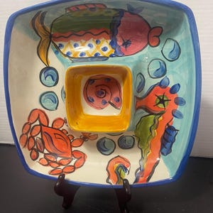 Op de afbeelding: Een vierkante keramische serveerschaal met een grillig ontwerp van het zeeleven. De schaal heeft een centrale vierkante kom, omgeven door drie secties, elk versierd met kleurrijke illustraties van een vis, een krab en een zeepaardje. De rand is blauw en de schaal wordt ondersteund door een bruine standaard.